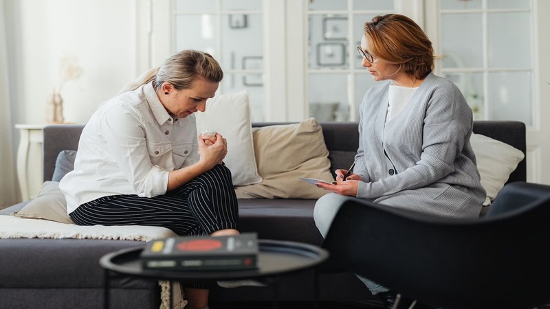 Two people sitting on a couch, one holding a clipboard, possibly in a counseling session. A small table with books is in the foreground.